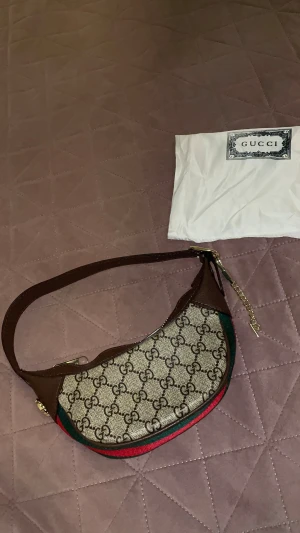 Gucci axelväska med GG-mönster - Snygg axelväska från Gucci med klassiskt GG-mönster i beige och brunt. Väskan har bruna skinnpartier, guldfärgade detaljer och dragkedja. Röd och grön rand längs kanten ger den en ikonisk Gucci-look. Perfekt storlek för det viktigaste och levereras med dustbag.