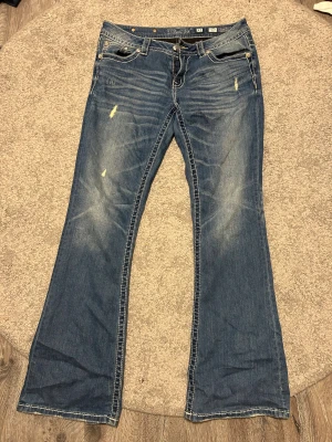 Miss Me Signature Bootcut jeans blå - Snygga blå jeans från Miss Me i modellen Signature Bootcut, med dekorativa sömmar och blingdetaljer på fickorna. Jeansen har klassisk femficksdesign, slitningar och kontraststickningar. Perfekt för dig som gillar en cool och avslappnad stil.