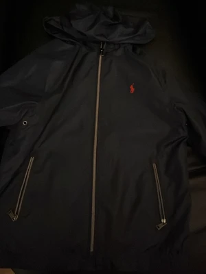 Mörkblå vindjacka från Polo Ralph Lauren - Säljer en mörkblå vindjacka från Polo Ralph Lauren med huva och dragkedja framtill. Jackan har två fickor med dragkedja och en röd broderad logga på bröstet. Perfekt för blåsiga dagar och enkel att matcha med det mesta.