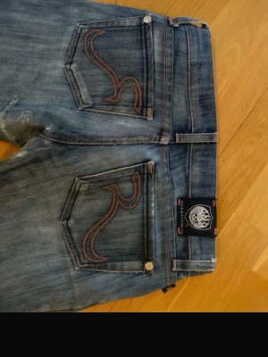 Rock & Republic blå bootcut jeans - Snygga blå jeans från Rock & Republic med bootcut passform och låg midja. Jeansen har slitna detaljer och rosa broderi. Materialet är jeans med en tvättad look.