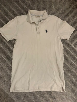  pikétröja från U.S. Polo Assn. - Snygg vit (kräm vit) pikétröja från U.S. Polo Assn i storlek S. med klassisk krage och korta ärmar. Tröjan har knappar vid halsen och en mörkblå broderad logga på bröstet. Perfekt för en chill och stilren look.