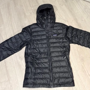 Svart dunjacka från Patagonia - Säljer en svart dunjacka från Patagonia med huva och dragkedja framtill. Jackan har två sidofickor med dragkedja och quiltad design. Perfekt för kyliga dagar och enkel att matcha med allt. Pufferjacka i storlek S.