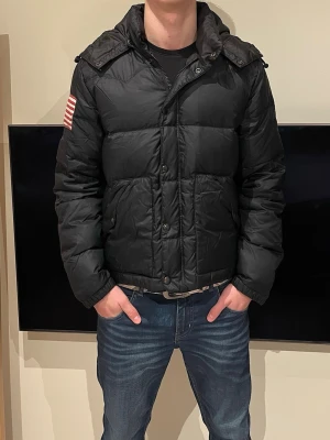 Svart pufferjacka från Ralph Lauren - Svart pufferjacka från Ralph Lauren med huva och amerikansk flagga på vänster ärm. Jackan har stora fickor med knappar och är tillverkad i polyester. Perfekt för kalla dagar och har en schysst form.