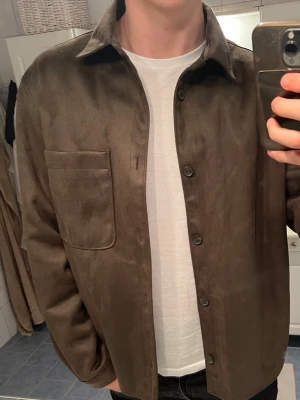 Brun mocka overshirt från Zara - Säljer en brun overshirt från Zara i storlek L. Jackan är gjord i mjuk mocka-look med klassisk krage, stora knappar framtill och en stor bröstficka. Perfekt för lager-på-lager och har en clean, stilren vibe.