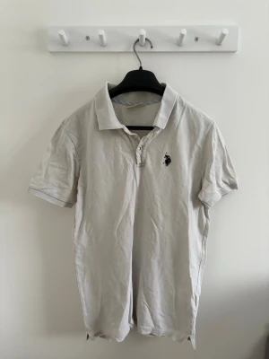 Herr kräm kortärmad pikétröja  - Snygg vit (kräm vit) pikétröja från U.S. Polo Assn i storlek S. med klassisk krage och korta ärmar. Tröjan har knappar vid halsen och en mörkblå broderad logga på bröstet. Perfekt för en chill och stilren look. 