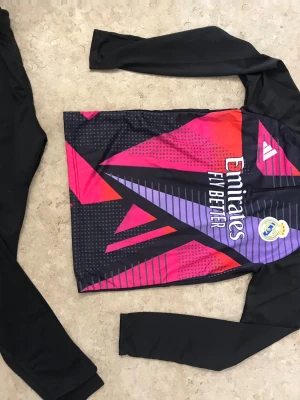 Real Madrid träningströja Adidas - Säljer en långärmad Real Madrid träningströja från Adidas med grafiskt mönster i svart, lila och rött. Tröjan har klubbmärke och sponsortryck på bröstet samt en sportig design. Perfekt för dig som älskar fotboll och vill träna i stil.