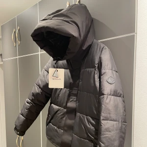 Moncler jacka  - Säljer en svart pufferjacka från Moncler med stor huva och diskret logga på ärmen. Jackan har dragkedja och knappar framtill samt breda, quiltade paneler. Perfekt för kalla dagar och riktigt snygg till streetwear-stilen.