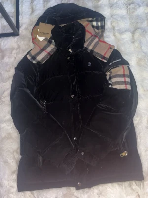 Svart pufferjacka från Burberry S - Säljer en svart pufferjacka från Burberry i storlek S. Jackan har klassiskt rutigt foder och detaljer i beige, rött och svart. Ikonisk Burberry-logga på bröstet och axeln, samt dragkedja och knappar framtill. Perfekt för kalla dagar och riktigt snygg streetstil.