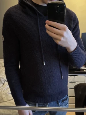 Mörkblå mocler hoodie - Säljer en mörkblå stickad hoodie från Moncler med huva och snörning. Tröjan har långärmade ribbade muddar och Moncler-logga på ärmen. Perfekt för dig som gillar exklusiva märken och vill ha en chill look.