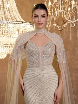 glittrig långklänning - Magisk långklänning i champagnefärg med glittriga detaljer och pärlor. Klänningen har en figurnära passform, transparent cape med fransar och hög krage. Materialet är skimrande och elegant, perfekt för bal eller fest. Djup v-ringning framtill och öppen rygg ger en lyxig känsla.