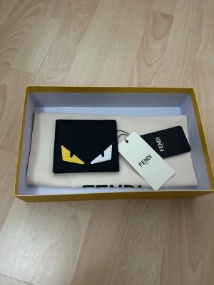 Svart plånbok från Fendi  - Säljer en svart plånbok från Fendi i skinn med ikoniska gula och vita ögon på framsidan. Plånboken har flera fack för kort och ID, och kommer i original ask och dustbag. Perfekt för dig som vill ha något unikt och stilrent i fickan.