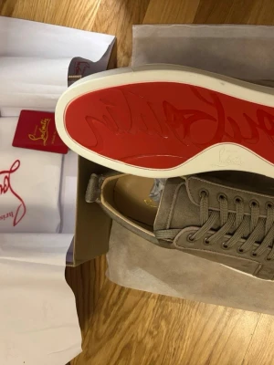 Christian Louboutin grå mockasneakers - Snygga sneakers från Christian Louboutin i grå mocka med klassisk röd sula och vita kanter. Skorna har rund tå, snörning och diskreta detaljer. Perfekta för dig som vill ha något stilrent men ändå sticker ut med den ikoniska röda sulan.