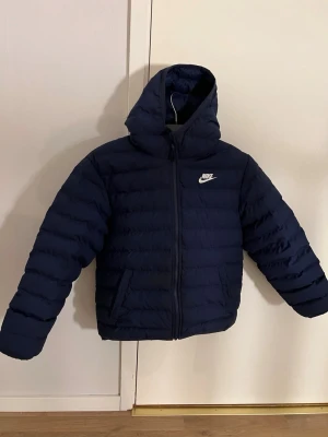 Mörkblå pufferjacka från Nike - Säljer en mörkblå pufferjacka från Nike med huva och dragkedja framtill. Jackan har ett stilrent, quiltat mönster och två fickor på sidorna. Perfekt för kalla dagar och riktigt skön att bära. Nike-logga i vitt på bröstet för en sportig vibe.