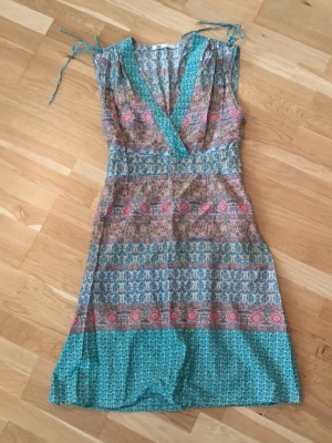 Blommig bohemisk klänning - En färgglad omlottklänning från Flash i storlek S. Klänningen har blommigt mönster i turkos, rosa och beige, v-ringning och knytband på axlarna.