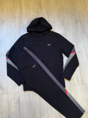 Under Armour tracksuit - Snyggt svart träningsset från Under Armour med hoodie och matchande byxor. Setet har grå och röda detaljer längs ärmar och ben, samt diskret logga på bröstet och benet. Materialet är mjukt och syntetiskt, perfekt för träning eller chill. Byxorna har dragsko i midjan.