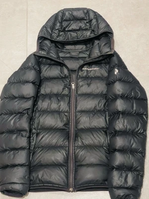 Peakperformance jacka - Svart dunjacka från Peak Performance i storlek S. Jackan har huva, quiltad design och dragkedja framtill. Perfekt för kalla dagar och har en clean look som funkar till många stilar. Materialet är syntet och dun, vilket gör den både lätt och varm.