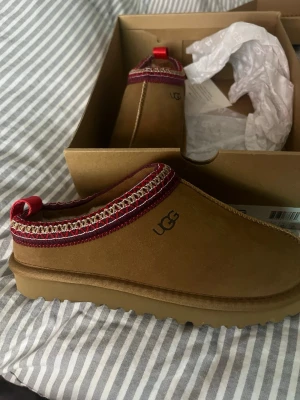 Oanvända UGGs - Bruna UGGs i mjuk mocka med fluffigt ullfoder. Detaljer längs kanten i vinrött och beige med broderade detaljer röd hälflik. Perfekta för kalla dagar och chill hemma.   STORLEK 38, Helt oanvända och pris kan diskuteras!