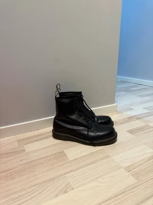 Svarta Dr. Martens boots strl 45 - Klassiska svarta Dr. Martens boots i skinn med gul söm runt sulan och dragflik bak. Modellen har snörning och rund tå, perfekt för dig som vill ha en tidlös och edgy look. Storlek EU 45, UK 10, US 11. Knappt använda (fick de som julklapp, men ändrade still snabbt efter)