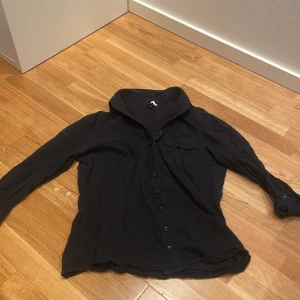 Svart skjorta med ficka och knappar - Svart skjorta i linnetyg med klassisk krage och tre kvart armar . Framtill finns en bröstficka med knapp och hela skjortan stängs med svarta knappar. Passformen är lite kroppad 