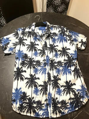 Vit och blå palmmönstrad skjorta - Kortärmad skjorta från Jack & Jones ( Gibraltar ) med palmmönster i svart och blått på vit botten. Skjortan har klassisk krage och knappar framtill, perfekt för sommarens chill dagar. 