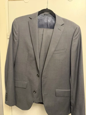 Grå smalrandig kostym från Riley - Säljer en stilren grå kostym från Riley i slim fit-modell. Kavajen har tunna vita ränder, två knappar framtill och klassiska fickor med lock. Tillverkad i ett mjukt material som känns bekvämt och passar perfekt till mer uppklädda tillfällen. 