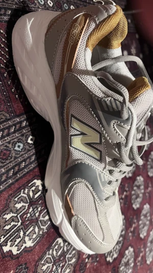 New Balance 530 sneakers grå/guld - Säljer ett par New Balance 530 sneakers i grått och guld med vita detaljer. Skorna har mesh och syntetpaneler, klassisk snörning och en chunky vit sula. Perfekta för dig som gillar retrostil och vill ha bekväma sneakers till vardags.