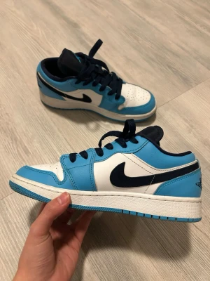Nike Air Jordan 1 Low blå/vit - Säljer ett par Nike Air Jordan 1 Low i fräscha blå och vita färger med svarta detaljer. Skorna har klassisk snörning, rund tå och platt sula. Materialet är skinn och textil, med den ikoniska Jumpman-loggan på tungan. Perfekt för dig som gillar streetwear och sneakers.