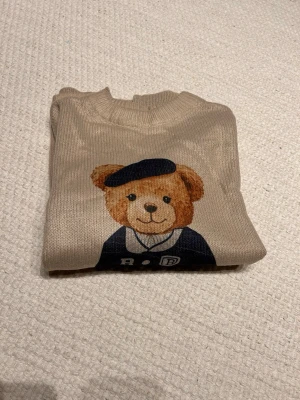 Beige stickad tröja med björnmotiv - Beige stickad tröja med rund hals och lång ärm. På framsidan finns ett stort tryck av en söt björn i marinblå outfit och basker. Tröjan har ribbade muddar och en avslappnad passform. Perfekt för dig som gillar gulliga detaljer och vill ha något unikt i garderoben.