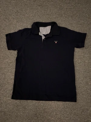 Blå pikétröja från GANT - Snygg blå pikétröja från GANT med klassisk krage och korta ärmar. Tröjan har en liten broderad logga på bröstet och insida krage med diskret randigt tyg. Perfekt för en chill och stilren look.