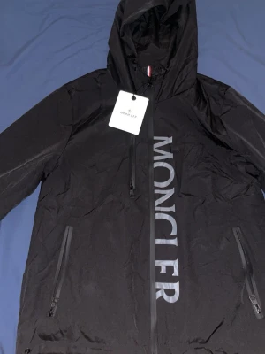 Svart vindjacka från Moncler - Svart vindjacka från Moncler med stor logga framtill. Jackan har huva, flera dragkedjeförsedda fickor och snygga detaljer. Materialet är lätt och känns sportigt, perfekt för blåsiga dagar. Klassisk passform och stilren look med diskret glans.