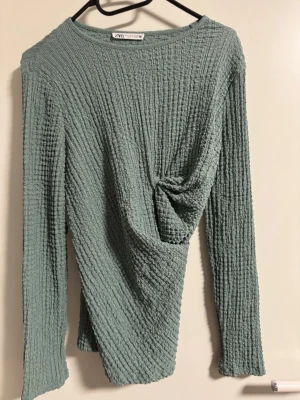 Grön ribbad topp från Zara - Säljer en grön långärmad topp från Zara med ribbad struktur och snygg twistdetalj framtill. Toppen har en mjuk, vågig textur och en avslappnad passform som ger en cool look. Perfekt att styla med jeans eller kjol.