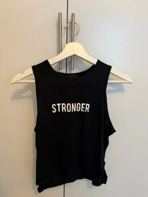 Svart cropped träningstopp Stronger - Säljer en svart cropped träningstopp från Stronger med vit text framtill. Tillverkad i 90% återvunnen polyester och 10% elastan för en stretchig och bekväm känsla. Perfekt för gymmet eller andra träningspass.