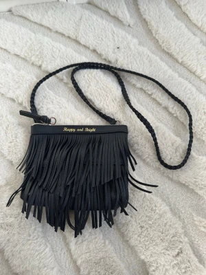 Svart axelväska med fransar - Säljer en svart axelväska  med långa fransar framtill och flätat axelband. Väskan är i skinnimitation och har dragkedja upptill. Perfekt för dig som vill ha en cool och unik accessoar.