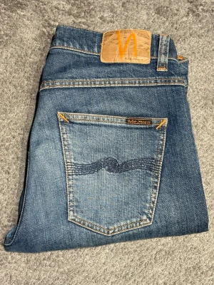 Nudie Jeans blå jeans 31/34 - Säljer ett par klassiska blå jeans från Nudie Jeans i storlek 31/34. Jeansen har raka ben, fem fickor och snygga kontrastsömmar samt Nudie-logga på bakfickan. Tillverkade i slitstark bomull med en tvättad finish för en avslappnad look.