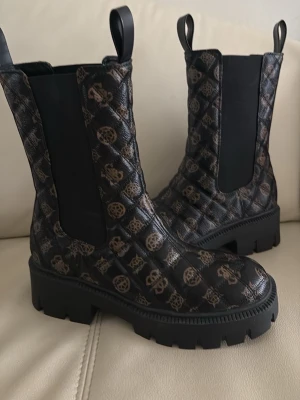 Svarta  boots från Guess - Säljer ett par svarta Chelsea boots från Guess med chunky sula och brunt logomönster över hela skon.