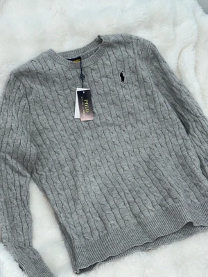 Grå kabelstickad tröja Polo Ralph Lauren - Säljer en grå kabelstickad tröja från Polo Ralph Lauren med rund halsringning och diskret broderad logga på bröstet. Tröjan har långa ärmar och ribbade muddar vid ärmslut och nederkant. Perfekt för lager på lager under kyliga dagar.