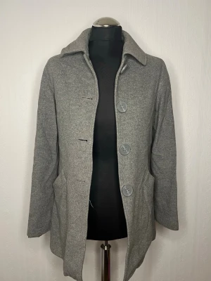 Grå från MNG XS - Säljer en stilren grå kappa från MNG i storlek XS. Kappan har stora knappar framtill, två fickor och klassisk krage. Enkel och clean design som passar till allt.