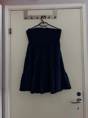 Marinblå axelbandslös kortklänning - Säljer en marinblå axelbandslös kortklänning med smock upptill och ett lätt utställd kjol. Klänningen har en enkel, stilren look och är gjord i ett mjukt, strukturerat tyg som ger en skön känsla. Perfekt för varma dagar eller när du vill ha något bekvämt och snyggt.