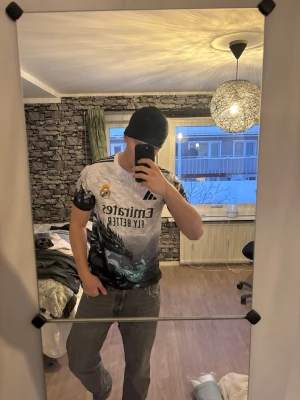 Real Madrid x Adidas draktröja - Säljer en unik Real Madrid t-shirt från Adidas med ett fett drakmotiv i svart, vitt och turkost. Tröjan har korta ärmar, klassiska Adidas-ränder på axlarna och Emirates Fly Better-tryck på bröstet. Perfekt för dig som vill sticka ut på läktaren eller i vardagen.
