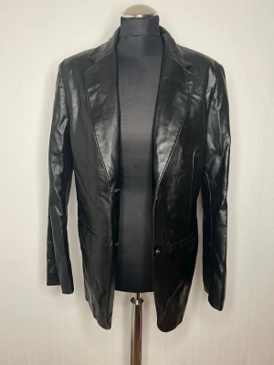 Svart skinnkavaj från H&M XS - Säljer en svart kavaj i skinnimitation från H&M i storlek XS. Kavajen har klassisk krage, två knappar framtill och fickdetaljer. Snygg och stilren modell som ger en edgy vibe till din outfit. Legat nerpackad i förrådet och aldrig blivit använd… 