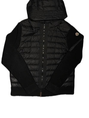 Svart Moncler pufferjacka med huva - Säljer en svart pufferjacka från Moncler med stickade ärmar och quiltad kropp. Jackan har dragkedja framtill, huva och en diskret Moncler-logga på ärmen. Perfekt för kyliga dagar och riktigt snygg till streetwear-stilen.