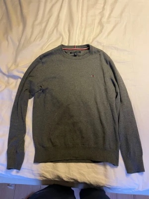  Stickad tröja Tommy Hilfiger  - Snygg grå stickad tröja från Tommy Hilfiger i storlek XS. Klassisk rund hals med ribbade muddar och diskret logga broderad på bröstet. Perfekt för dig som gillar stilrena och enkla plagg med en touch av preppy. Passar dig som vill ha en normal passform. 