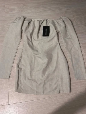 Beige offshoulder klänning PLT - Trendig beige kortklänning från PrettyLittleThing med offshoulder-design och långa ärmar. Klänningen har en liten slits nertill och är gjord i ett lätt, vävt material som känns luftigt. Perfekt för dig som gillar stilrena och enkla plagg med en twist.