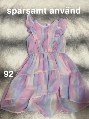 Pastellfärgad volangklänning barn - Supersöt klänning i pastellfärger med volanger. Klänningen har ett luftigt, lager-på-lager tyg i rosa, lila och blå toner. Ärmlös modell med volangdetaljer på axlarna och elastisk midja för skön passform.🌸 Sparsamt använd. Storlek 92