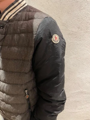 Moncler jacka  - Säljer en snygg bomberjacka från Moncler med quiltad grå front och rygg i ullmix och svarta ärmar i polyester. Jackan har randig ribbad krage och muddar, samt Moncler-logga på ärmen. Stängs med knappar och har dragkedjeförsedda fickor.