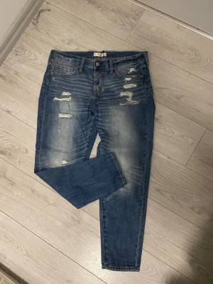 Blå jeans från Abercrombie & Fitch W30 - Säljer ett par blå jeans från Abercrombie & Fitch i storlek W30. Jeansen har slitna detaljer och snygga broderade bakfickor med mörkblå sömmar. Klassisk femficksmodell med knapp och dragkedja. Fy fon va grisch. 