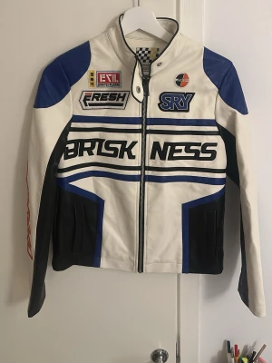 Vit och blå skinnjacka med racingtryck - Säljer en fet vit och blå skinnjacka med racinginspirerade tryck och patchar. Jackan har svarta detaljer, dragkedja framtill och coola grafiska linjer. Perfekt för dig som vill sticka ut med en sportig vibe. Snygg ståkrage och långärmad modell.