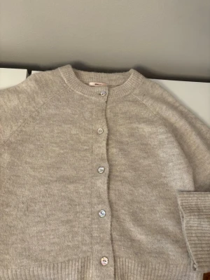 Beige kofta från Gina Tricot -  beige kofta från Gina Tricot i storlek 158/164. Koftan är stickad med ribbade muddar och har knappar framtill. Perfekt för lager på lager under kyliga dagar och enkel att matcha med allt.