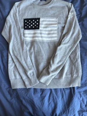 Grå stickad tröja från GANT med flagga - Säljer en grå stickad tröja från GANT i storlek M. Tröjan har en stor amerikansk flagga i vitt och svart på bröstet och ribbade muddar vid ärmslut och nederkant. Snygg detalj med broderad text på ena ärmen. Perfekt till höst och vinter!