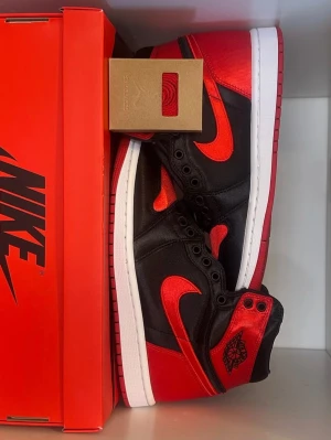 Nike Air Jordan 1 Satin Bred svart/röd - Nike Air Jordan 1 Satin Bred i svart och rött, i storlek 38 med Jordan Wings-logga vid hälen. Skorna har en blank satin-finish, röda swoosh-detaljer och svart snörning. Vit mellansula och röd yttersula. Svarta och röda skosnören kommer med. Säljer den för min lillebror som fick skorna för ungefär tre år sen men aldrig använts. Om du har några frågor så är det bara att skriva. Boxen ingår i köpet.